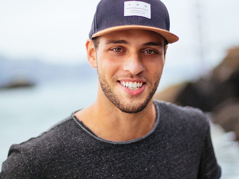 Smiling young man wearing a cap, standing outdoors near water, representing a friendly and approachable atmosphere for dental care services in Westchester County.