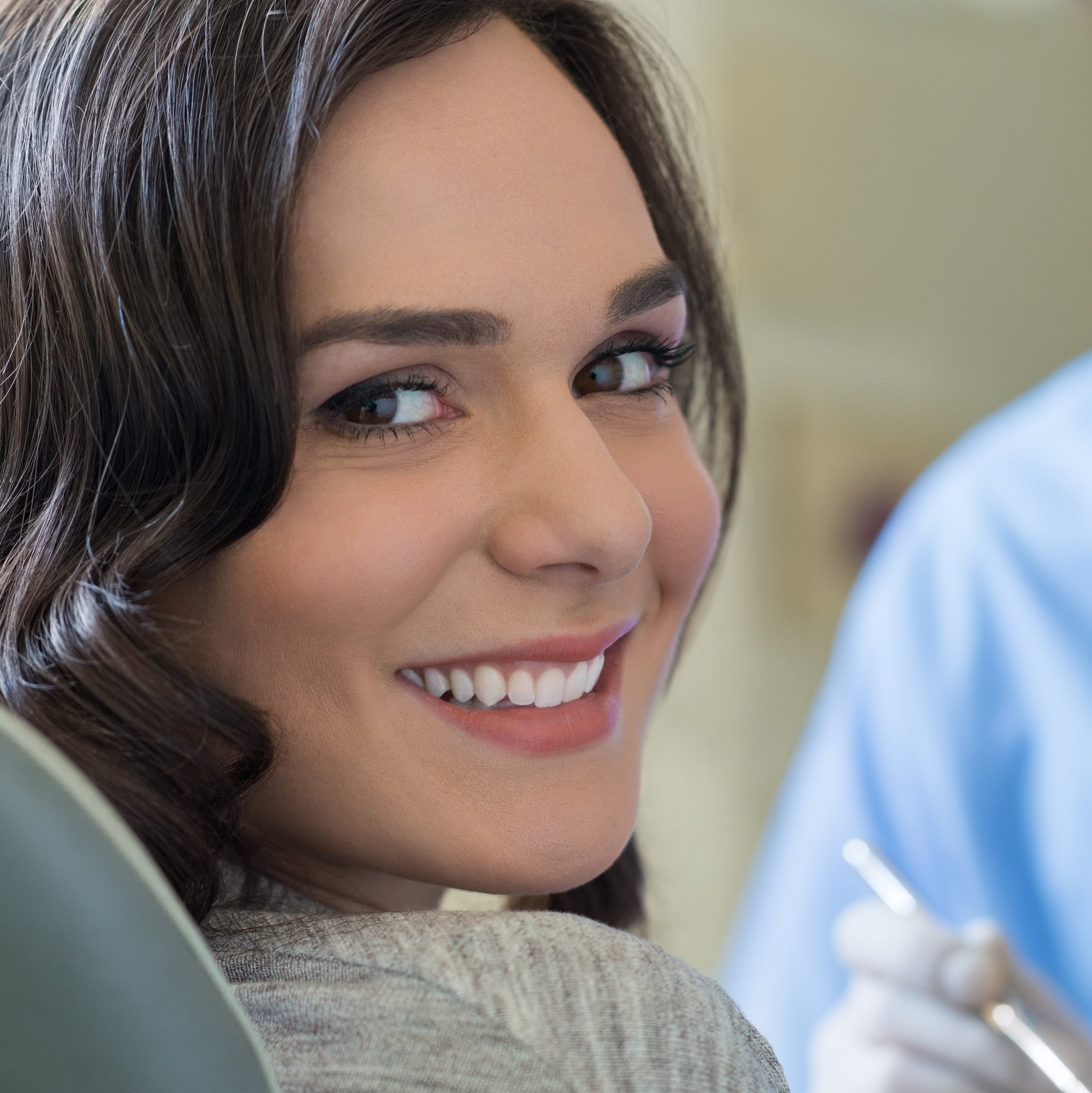Smiling patient in dental chair, showcasing a healthy smile, with dental professional in background preparing for treatment.