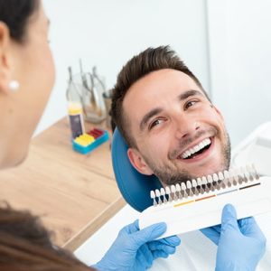 A man in dental chair while dental assistance shows different possible shades of teeth.